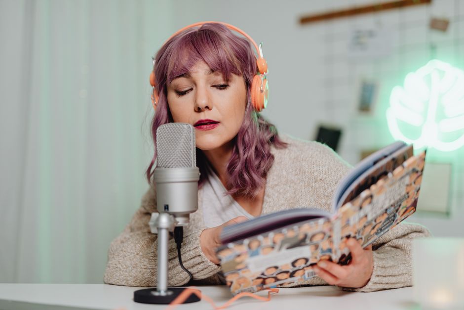 Woman with headphones reading a book into a microphone indoors.